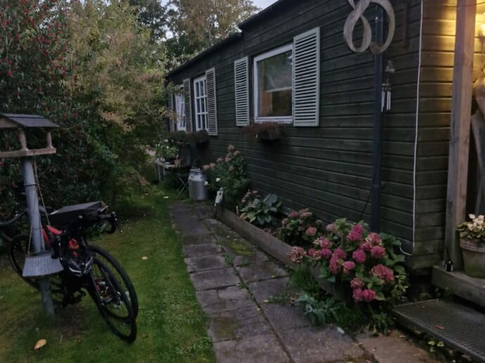 Et grønt træhus med nummer 85, med en stenbelagt sti, lyserøde hortensiabuske og en cykel, der står parkeret på græsset.